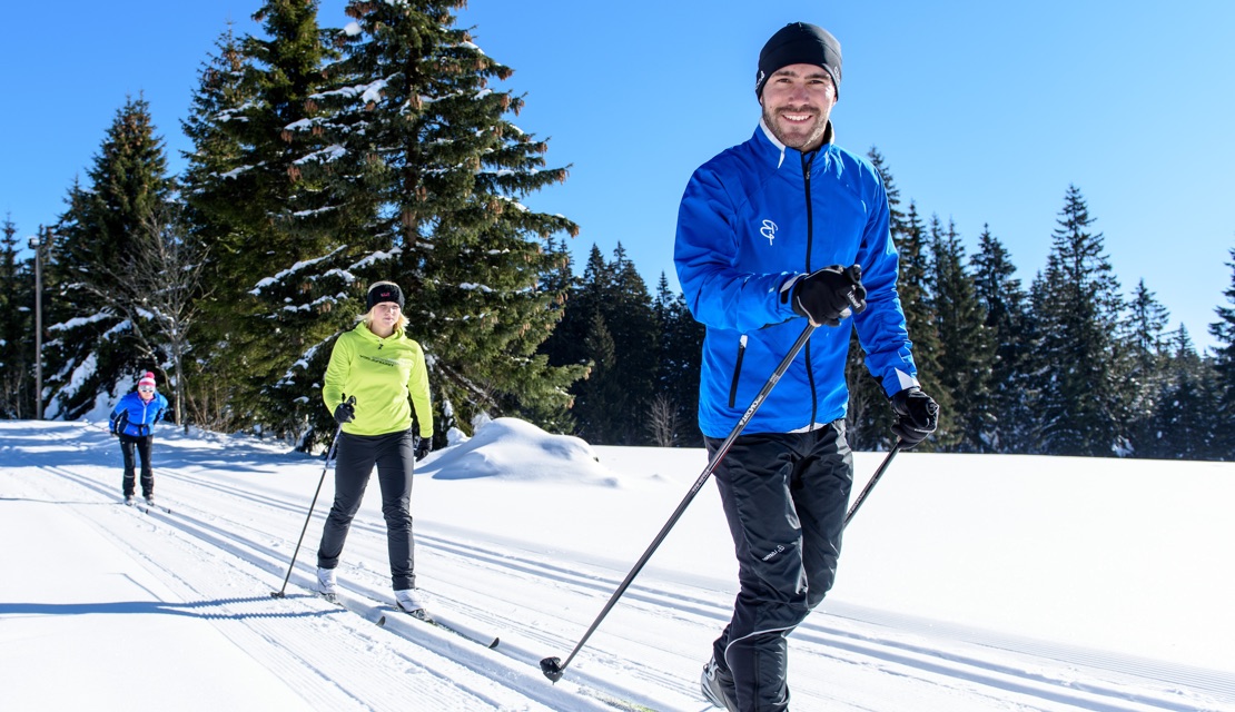 Drei Sportler machen klassischen Langlauf im Winter bei Schnee und mit Bäumen im Hintergrund