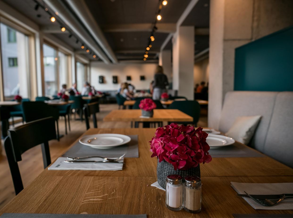 Eingedeckter Tisch im Restaurant Feinwerk mit roten Blumen im Vordergrund vor verschwommenem Hintergrund des belebten Restaurants